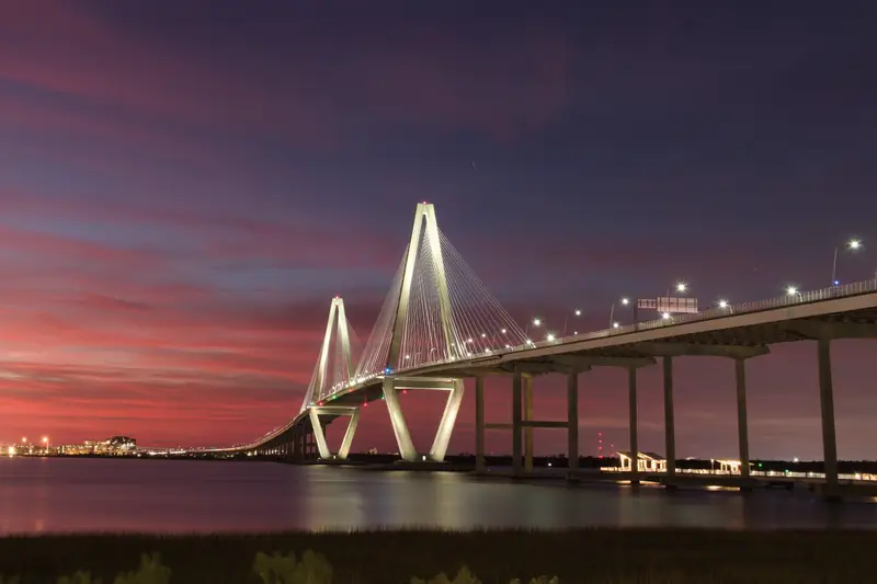Ponte estaiada iluminada sobre a água ao entardecer, com o céu rosa e roxo, em Charleston